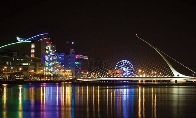 Photograph of Dublin city at night with the convention centre, Samuel Beckett bridge and a Ferris wheel in the background and a river in the foreground