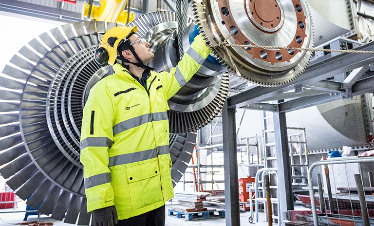 Man wearing high vis jacket and hard hat reaches up to fix a large piece of metal machinery