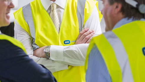 Close up photograph of a mans chest. He is crossing his arms and is wearing a high vis vest on top of a shirt and tie