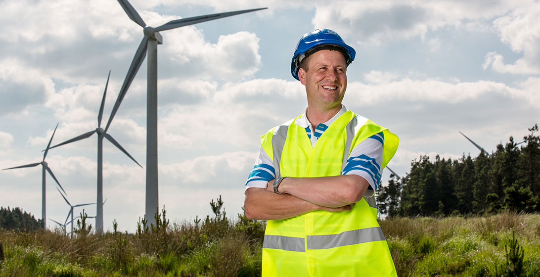 Photograph of a man standing in front of a windfarm. He is smiling and has his arms crossed. He is wearing a high vis vest and a hard hat