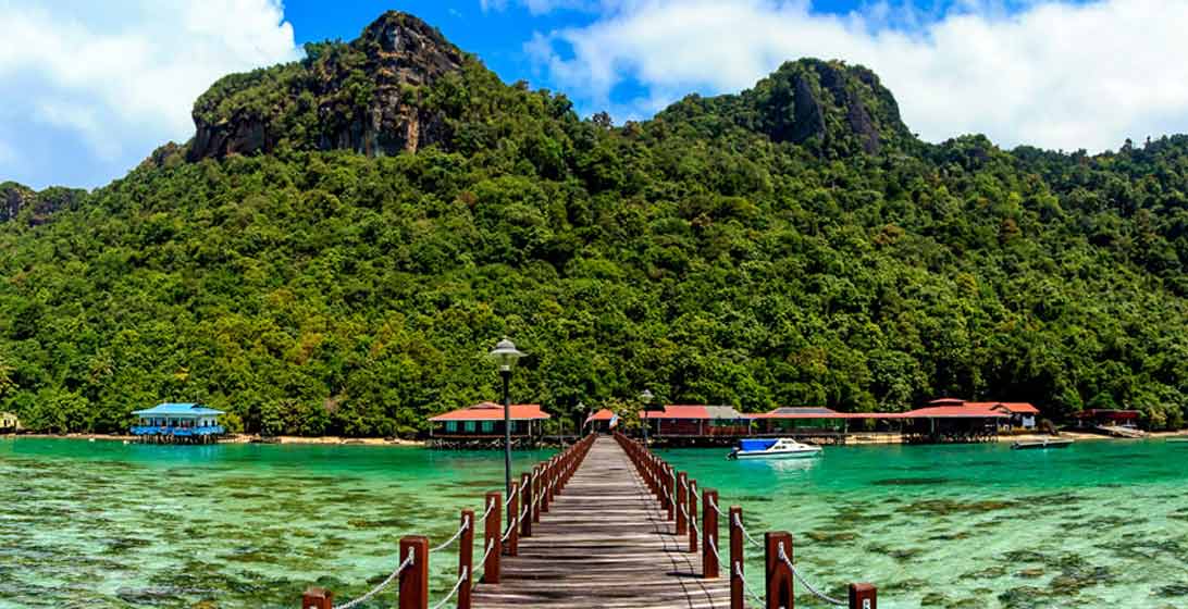 Photograph of a bridge over a green body of water leading to a forest covered mountain