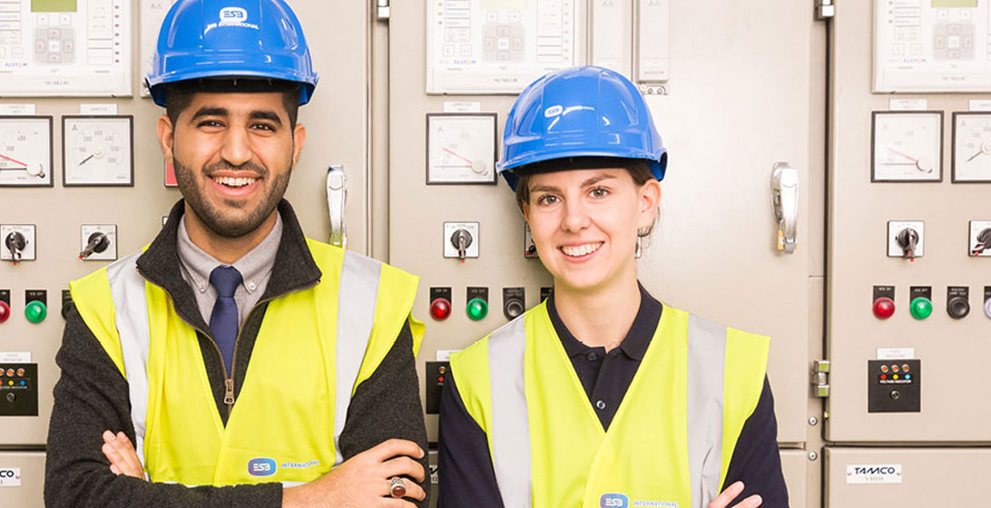 Photograph of a man and woman smiling and facing the camera. They are both wearing hard hats and high vis vests. Behind them are beige panels on the wall which contain many switches and gauges