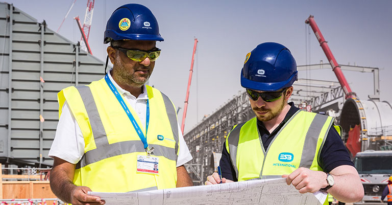 Image of two men in high vis vests and hard hats looking at some drawings on a building site