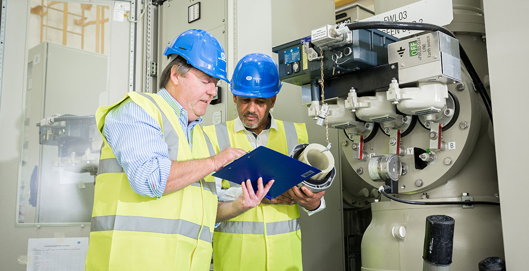 Photograph of two men in hard hats and high vis vests. They are looking at something on a clip board and standing next to some machinery
