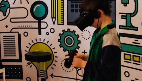 A photograph of a young boy using a virtual reality headset, standing in front of a monitor. There are illustrations on the wall behind him of technology objects