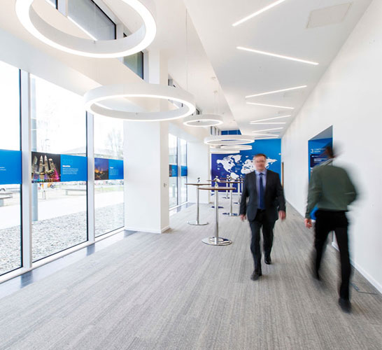 Image of two men walking past each other in a corridor with glazing on one wall and overhead lights