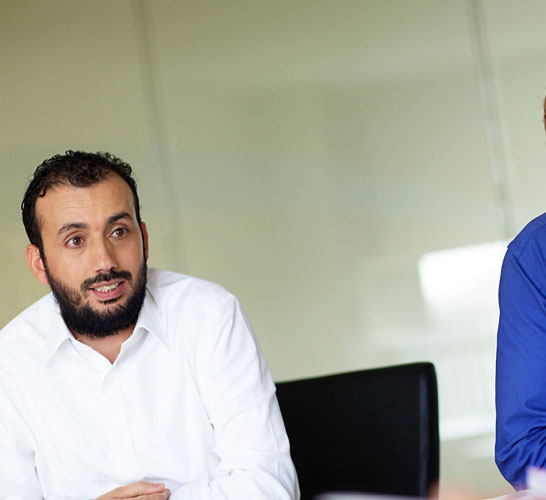 Image of two men in a meeting room listening to another man speak