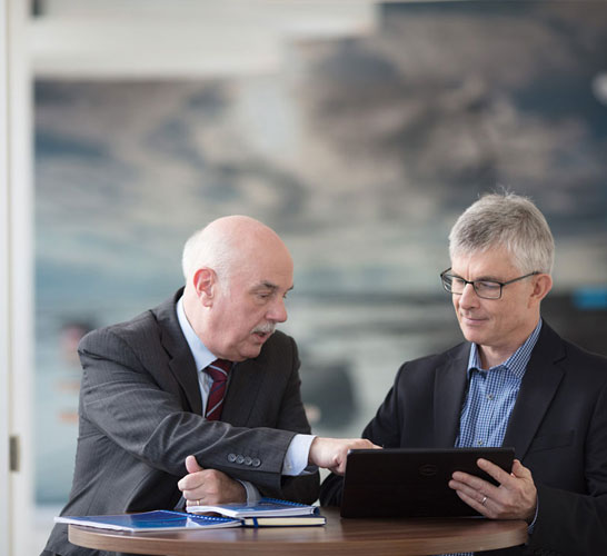 Image of two men in a meeting room listening to another man speak