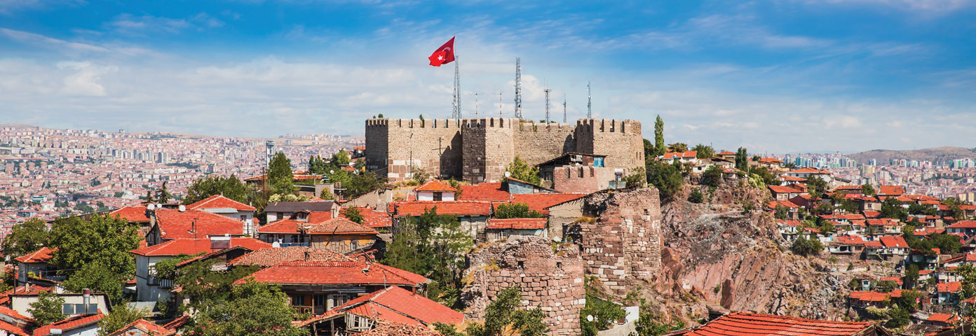 Photograph of a castle like structure at the top of a hill. It is surrounded by city and has a Turkish flag at the top