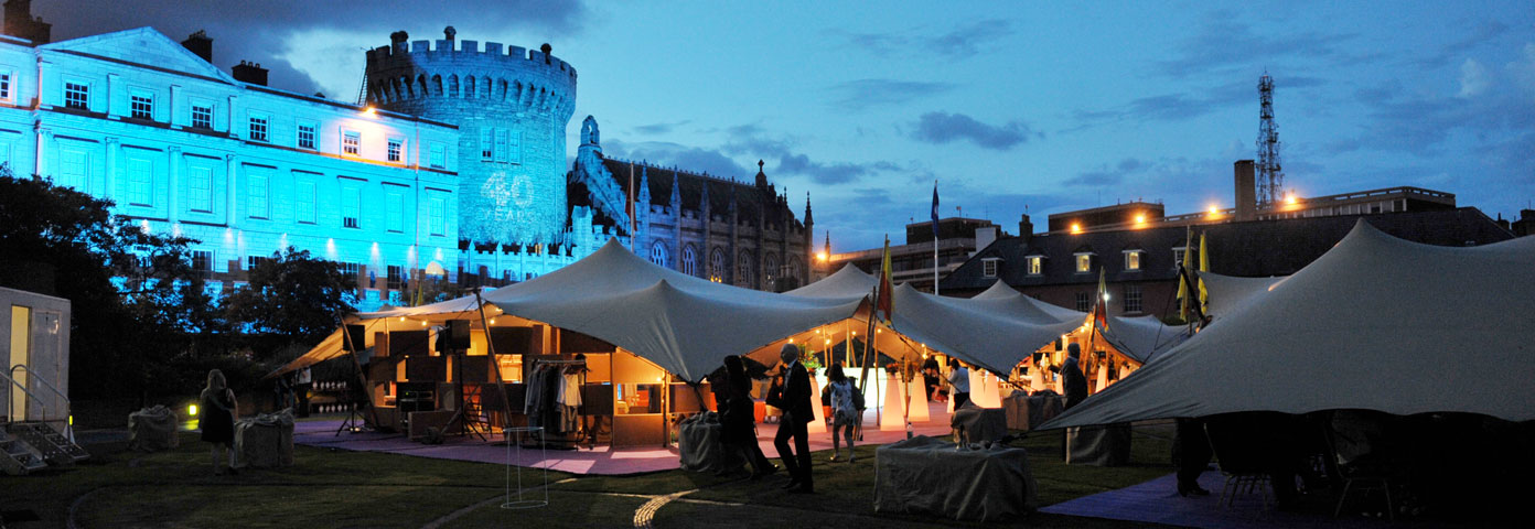Photograph of a castle lit up with blue lights. In front of it is a large tent with people and boxes inside