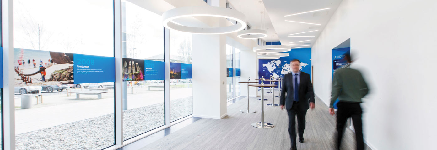 Image of two men walking past each other in a corridor with glazing on one wall and overhead lights